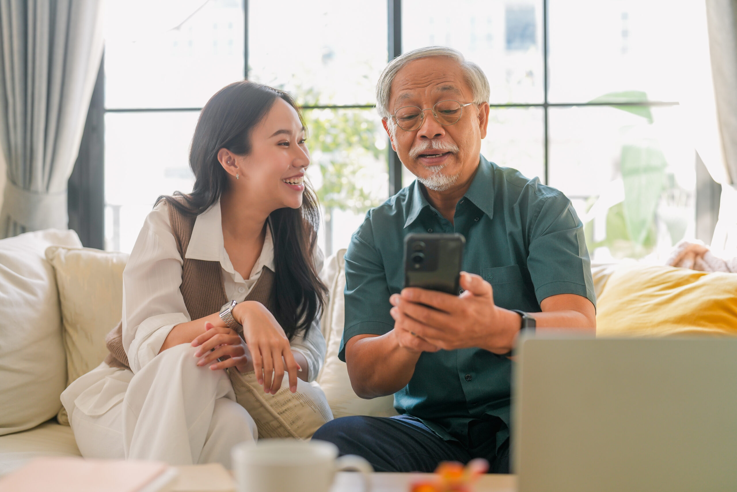 Lelaki warga emas dan cucunya duduk di sofa sambil melihat telefon dan ketawa berkaitan Kesihatan Digital