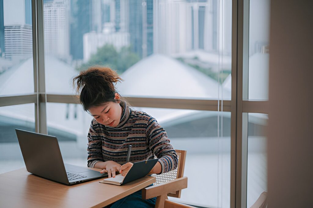 Female intern writing notes in a notebook next to a laptop on a desk by a large window